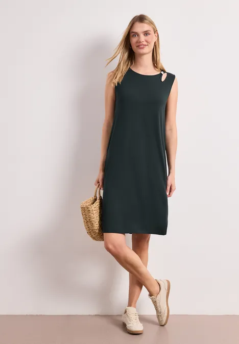 Dame in schlichtem schwarzem, ärmellosem Kleid mit One-Shoulder-Cut-out, Basttasche und weißen Sandaletten.