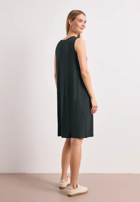 Eine Frau in schwarzem, knielangem, ärmellosem Kleid steht seitlich in Sneakers.