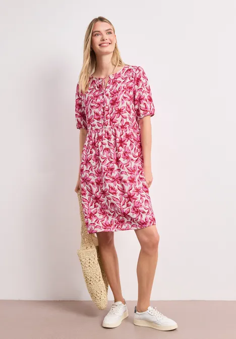 Femme en robe fluide rose à fleurs blanches, baskets blanches et sac en paille