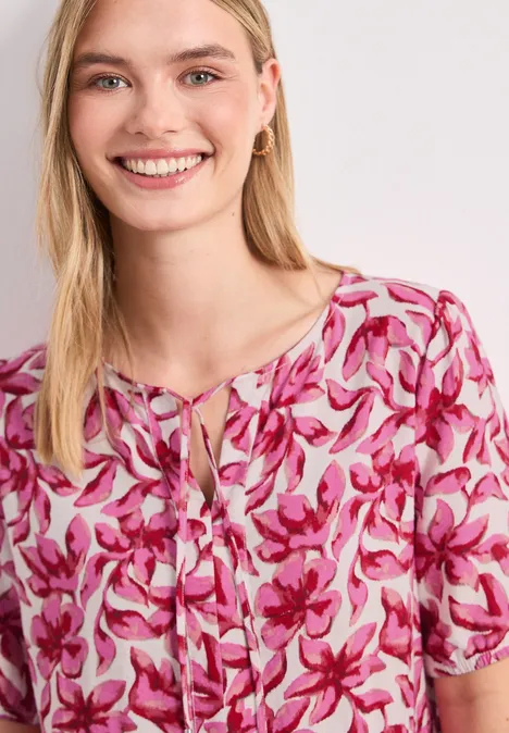 Femme portant un chemisier à fleurs rose et blanc, manches courtes et nœud décoratif au col.