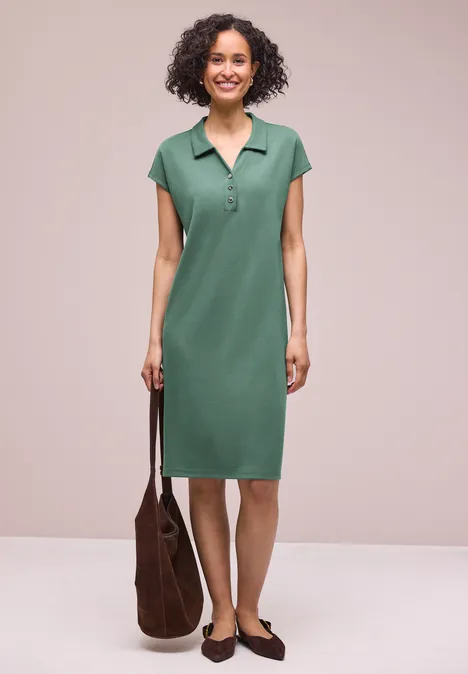 Frau in grünem Polokleid steht vor neutralem Hintergrund, trägt eine braune Tasche und Slipper.