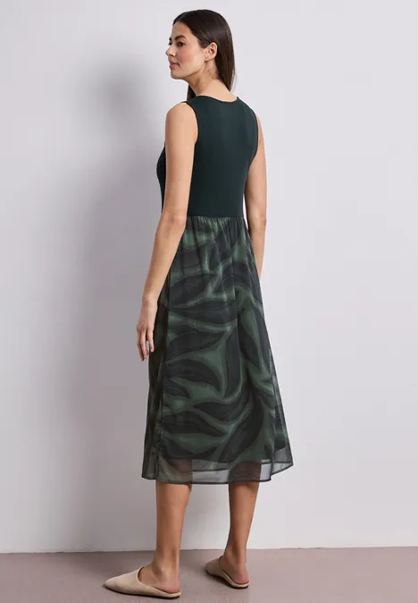 Vrouw van achteren in een donkergroen mouwloos shirt en groen gemêleerde midirok, lichte achtergrond.