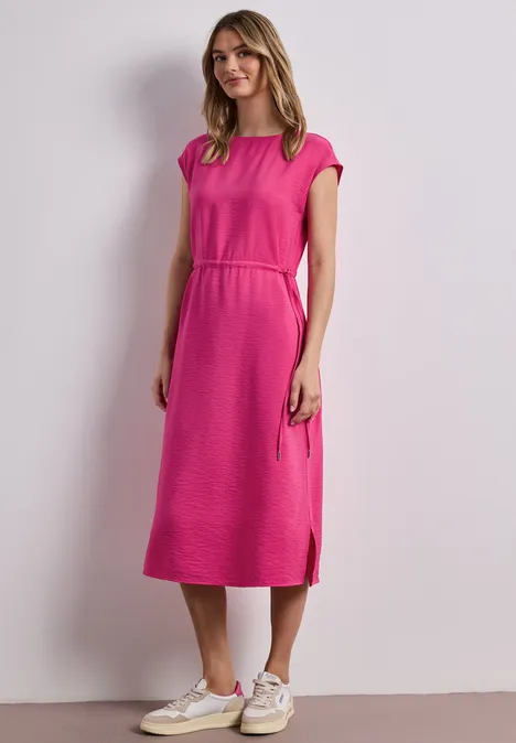 Kobieta w różowej sukience midi i białych trampkach na jasnym tle.