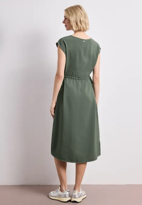 Eine Frau steht von hinten in einem ärmellosen, khakigrünen Midikleid mit weißen Sneakern vor neutralem Hintergrund.