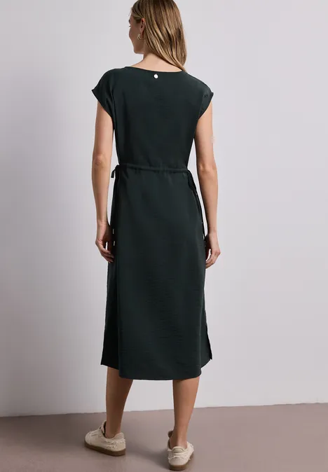 Frau von hinten in einem schlichten, midilangen schwarzen Kleid mit Taillengürtel und beigen Slippern.