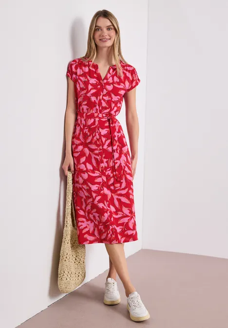 Frau trägt rotes Midikleid mit weißem Blattmuster, V-Ausschnitt, Taillengürtel und weiße Sneaker, hält groben Netzbeutel.