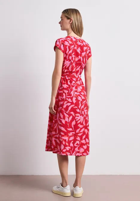 Frau im Profil trägt rotes Midikleid mit weißem Blattmuster und weiße Sneaker vor schlichtem Hintergrund.