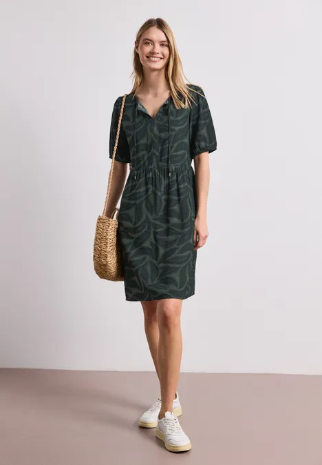Vrouw in donker groen overhemdjurk met bladprint, witte sneakers en gevlochten strooien tas.