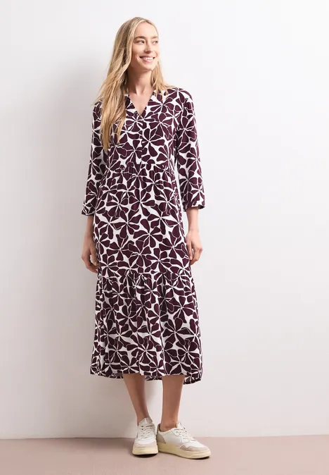 Midi-Kleid mit Split Neck und Print rustic rouge Blonde Frau trägt ein violett-weiß gemustertes Midikleid und weiße Sneaker vor weißem Hintergrund.