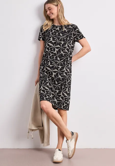 Vrouw in losvallende, knielange zwarte jurk met witte print, korte mouwen, witte sneakers en lichte jas.