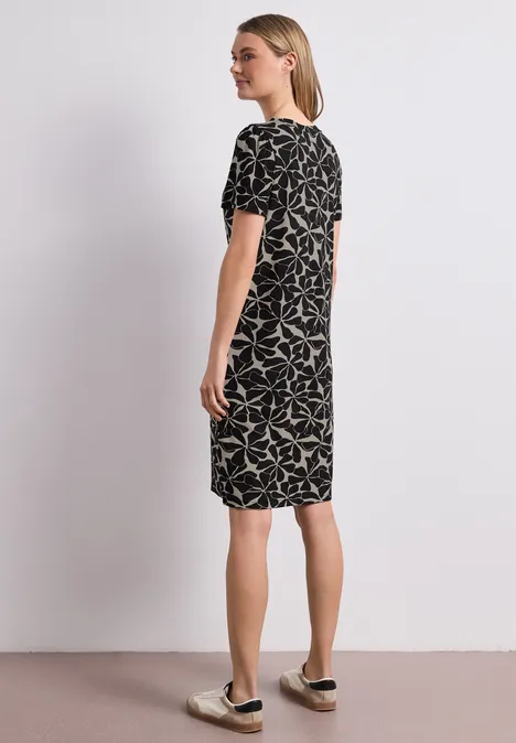 Vrouw staat in zijaanzicht tegen witte muur, draagt zwarte jurk met wit bladmotief en slip-on sneakers.