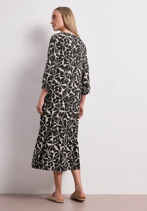 Midi-Kleid mit Split Neck im Ethno-Look Black Frau steht seitlich mit Blick nach hinten, trägt schwarzes Midikleid mit hellem Blattmuster und flache Sandalen.