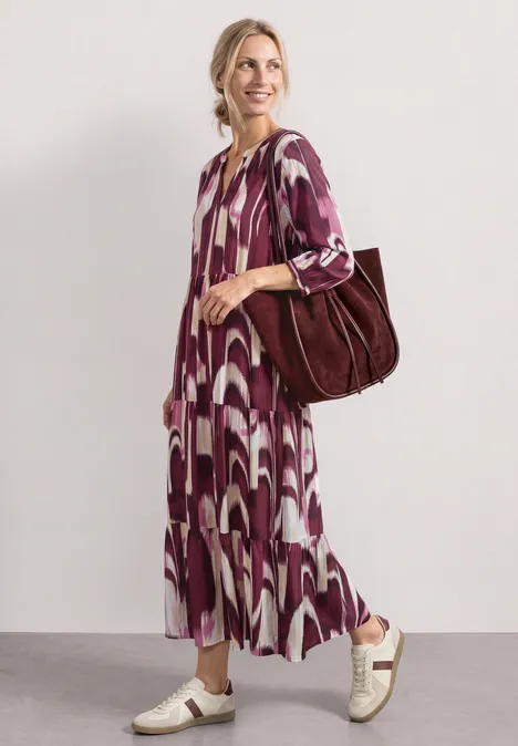 Vrouw in een midi-jurk met paarse-witte print, witte sneakers en grote bruine tas, loopt ontspannen.