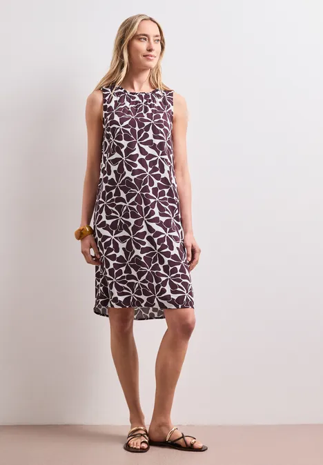 Knielange jurk met ronde halslijn rustic rouge Vrouw in mouwloze midi-jurk met bloemmotief en platte sandalen, staand voor een neutrale achtergrond