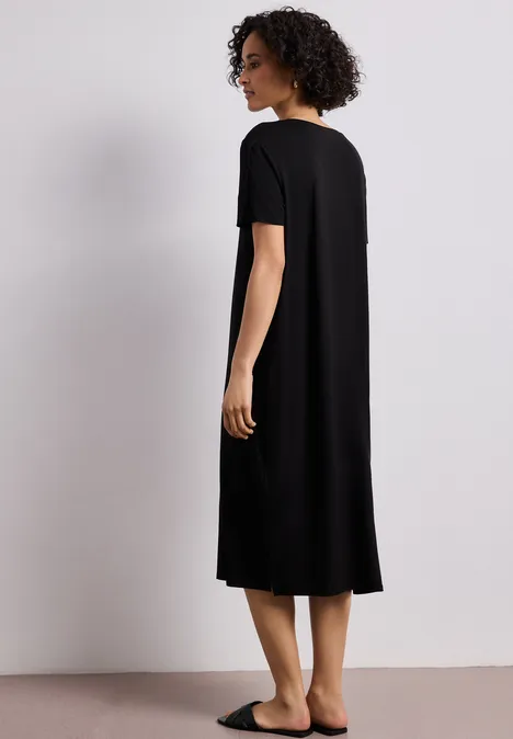 Midi-jurk met korte mouwen en splitdetail Black Een vrouw draagt een zwart, knielang T-shirtjurk en staat zijwaarts voor een neutrale achtergrond.