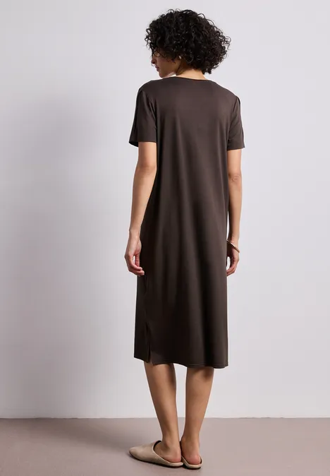 Eine Frau mit kurzem lockigem Haar steht vor neutralem Hintergrund, trägt ein schlichtes braunes Midikleid und Sandalen.