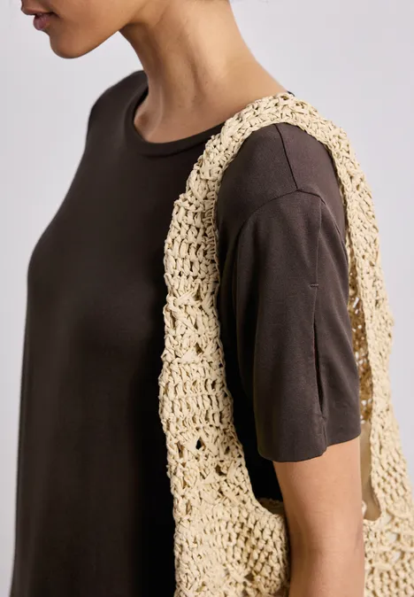 Person im dunkelbraunen T-Shirt mit gehäkelter beiger Tasche über der Schulter.