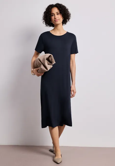 Frau trägt knielanges dunkelblaues T-Shirt-Kleid, hält beigefarbene Jacke, kombiniert mit flachen Schuhen.