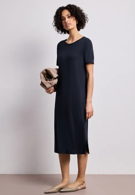 Midi-jurk met korte mouwen en splitdetail shadowed navy Vrouw in donkerblauw midi-jurk met korte mouwen en beige ballerina’s, houdt een licht vest vast.