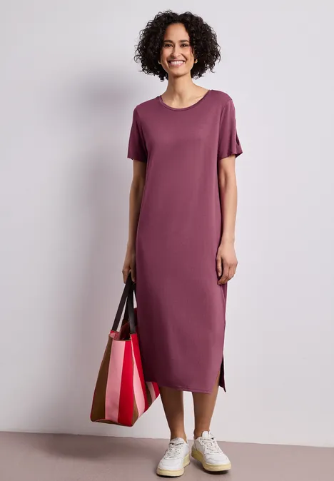 Person in lila Midi-T-Shirt-Kleid, weiße Sneaker und mehrfarbige Tragetasche.