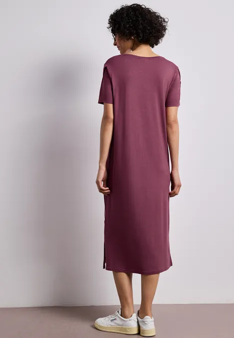 Midi-jurk met korte mouwen en splitdetail dark clouded blush Rugaanzicht van een vrouw in een simpel paars midijurk en witte sneakers tegen neutrale achtergrond.