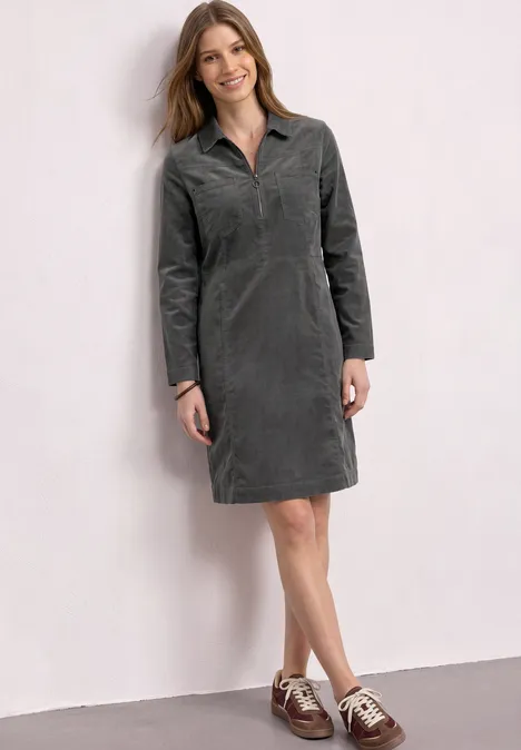 Kniehoge jurk van corduroy met ritssluiting gravel grey Vrouw in grijze corduroy jurk met kraag en lange mouwen en bruin-witte veterschoenen voor een witte muur.