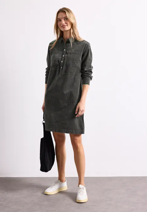 Knielanges Kleid aus softem Cord gravel grey Frau in dunkelgrünem Cord-Hemdkleid bis knapp über dem Knie, mit schwarzer Tasche und weißen Sneakern.