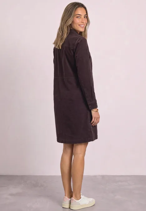Eine Frau steht seitlich, trägt ein knielanges, langärmeliges, dunkelbraunes Kleid und weiße Sneaker.