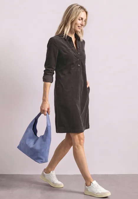 Eine junge Frau in dunkelgrauem Cordkleid und weißen Sneakern, die eine blaue Tasche trägt.