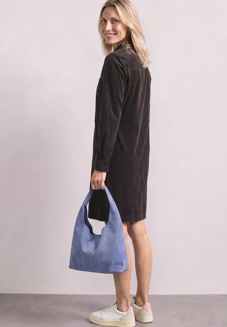 Frau in schwarzem Kleid und weißen Sneakers hält eine blaue Tasche und blickt lächelnd über die Schulter.