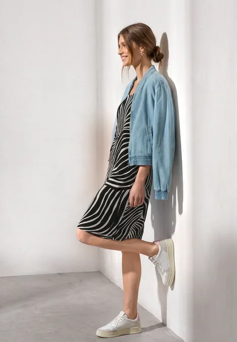 Eine Frau lehnt an einer weißen Wand und trägt ein Zebra-Midikleid, eine hellblaue Jeansjacke und weiße Sneaker.