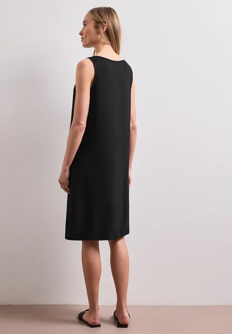 Knielanges Kleid mit Schnallendetail Black Frau von hinten in ärmellosem, knielangem schwarzen Kleid und flachen Sandalen vor neutralem Hintergrund.