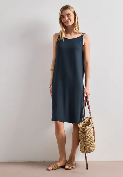 Knielanges Kleid mit Schnallendetail shadowed navy Frau in ärmellosem dunkelblauem Etuikleid mit Strohtasche und Sandalen vor schlichtem Hintergrund.