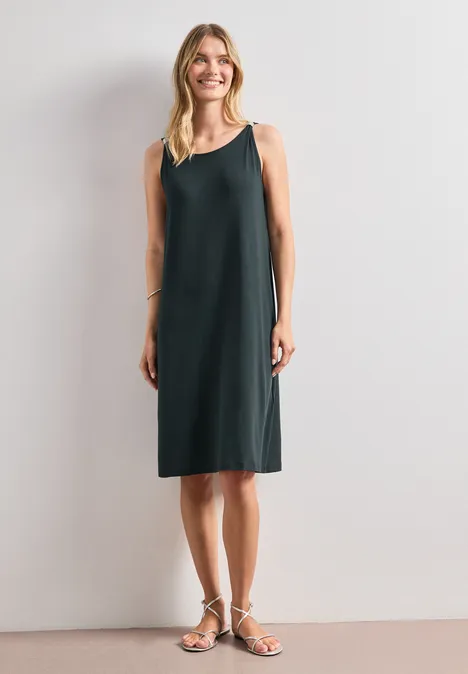 Robe longueur genou avec détail de boucle deep bound green Femme portant une robe vert foncé sans manches, longueur genou, et sandales, devant un mur clair.