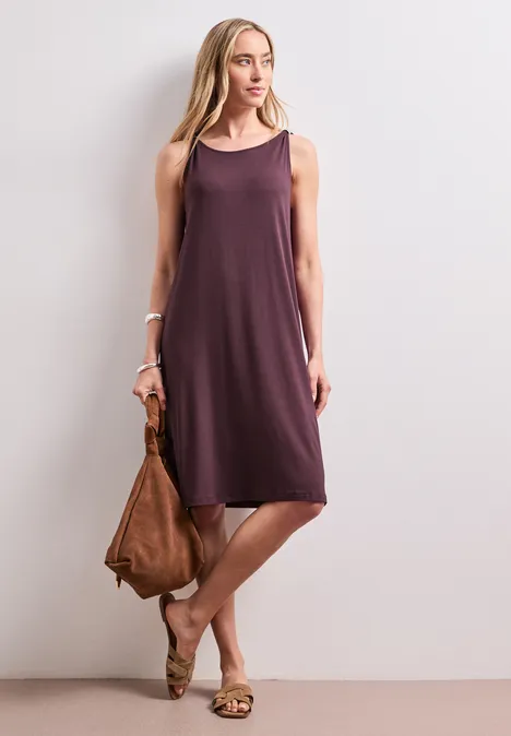 Knielanges Kleid mit Schnallendetail rustic rouge Frau in locker fallendem, ärmellosem violettem Kleid, mit brauner Tasche und Sandalen vor weißem Hintergrund.