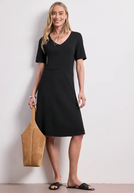 Knielanges Kleid aus Jersey mit Kurzarm Black Frau in schwarzem V-Ausschnitt-Kleid bis zum Knie, mit beiger Tragetasche und schwarzen Sandalen vor hellem Hintergrund.