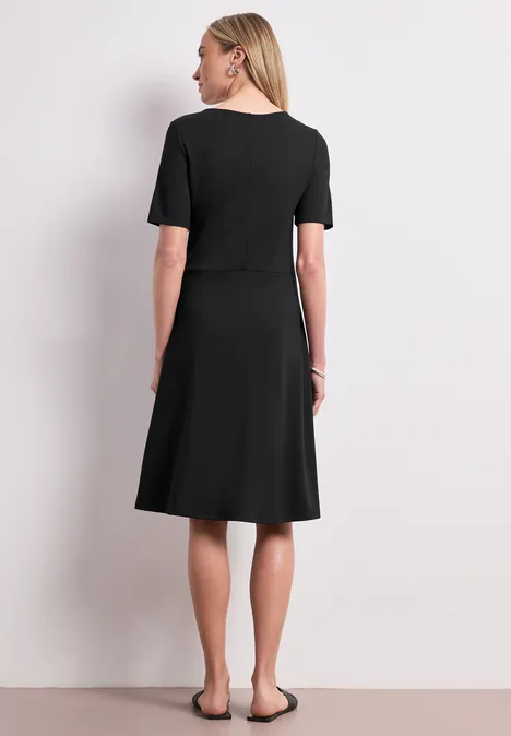 Knielanges Kleid aus Jersey mit Kurzarm Black Frau von hinten in schlichtem, knielangem, schwarzen Kleid mit kurzen Ärmeln und flachen Schuhen vor hellem Hintergrund.