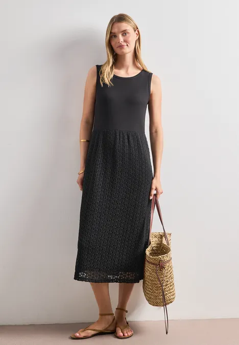 Frau in ärmellosem schwarzem Midikleid mit gehäkeltem Muster, Strohtasche und Sandalen vor weißem Hintergrund.