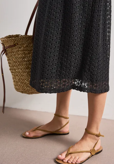 Midi-Kleid mit Rock aus Spitze Black Frau in schwarzem Häkelkleid mit Basttasche und flachen, olivgrünen, geschnürten Sandalen.