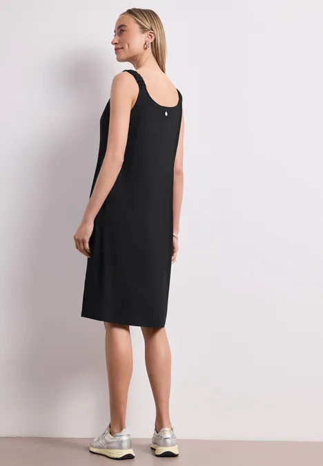 Knielanges Kleid mit gerafften Trägern Black Frau in schlichtem ärmellosem schwarzem Etuikleid knielang und weißen Sneakers, seitlich stehend vor hellem Hintergrund.