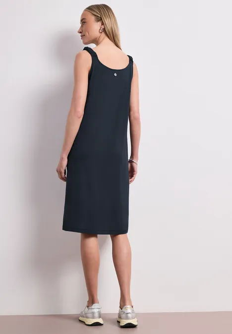 Knielanges Kleid mit gerafften Trägern shadowed navy Frau von hinten in ärmellosem, knielangem schwarzen Kleid und weißen Sneakern vor schlichter Wand.