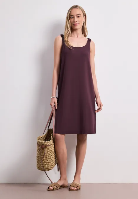 Knielanges Kleid mit gerafften Trägern rustic rouge Frau trägt ärmelloses, locker sitzendes lila Kleid, hält Strohtasche und Sandalen.