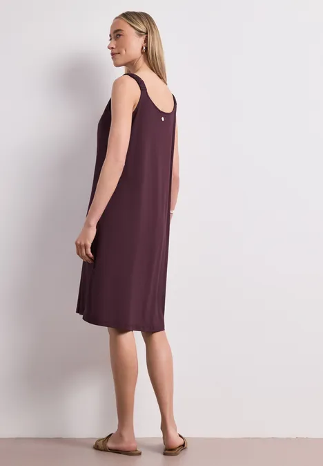 Knielanges Kleid mit gerafften Trägern rustic rouge Frau in lockerem, ärmellosem, knielangem, weinrotem Kleid und flachen Sandalen vor hellem Hintergrund