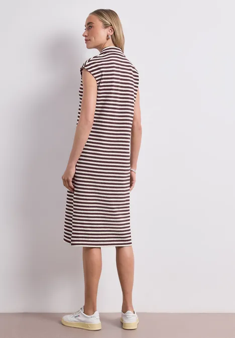 Gebreide midi-jurk met strepen rustic rouge Vrouw in een gestreepte knielange jurk en witte sneakers voor een lichtgrijze achtergrond.