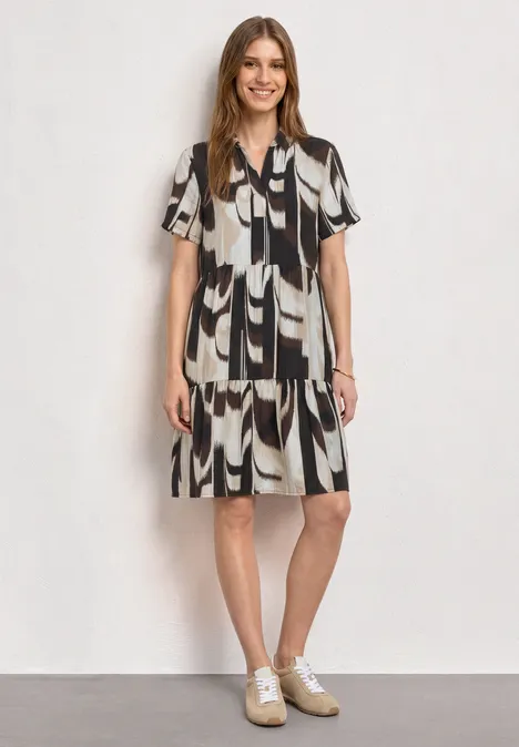 Vrouw in knielange zwart-wit jurk met abstract patroon en witte sneakers, tegen een lichte achtergrond.