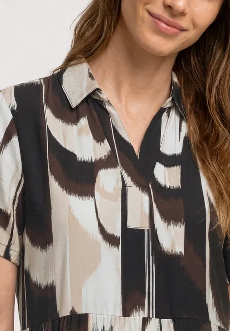 Dame draagt blouse met abstract penseelstreekpatroon in zwart, wit en beige, korte mouwen en V-hals.