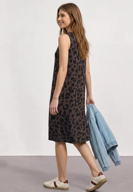 Frau in locker sitzendem, ärmellosem Animal-Print-Kleid, hält Jeansjacke und trägt Sneaker.