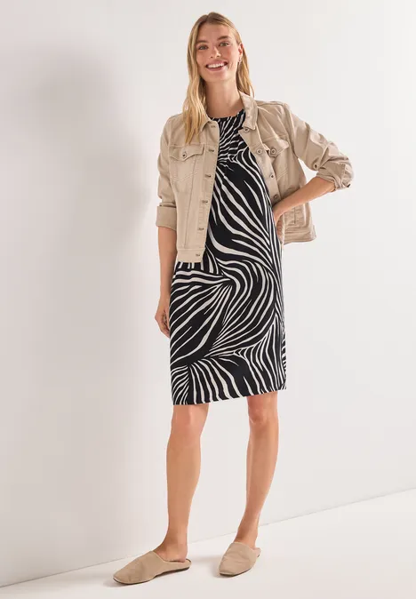 Frau in knielangem schwarz-weißem Zebra-Kleid, beige Jacke und bequemen Slippern.