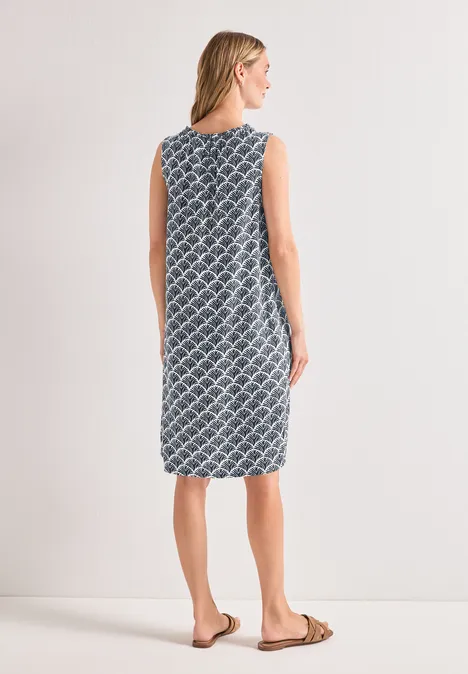Rückansicht einer Frau in ärmellosem, knielangem, blau-weißem, gemustertem Kleid mit flachen Sandalen.