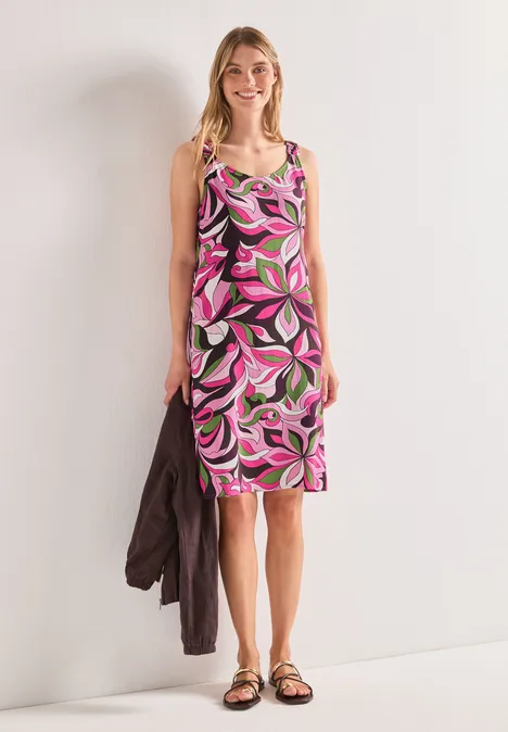 Femme en robe midi rose et verte à motifs, sandales, veste à l'épaule, devant un mur clair.
